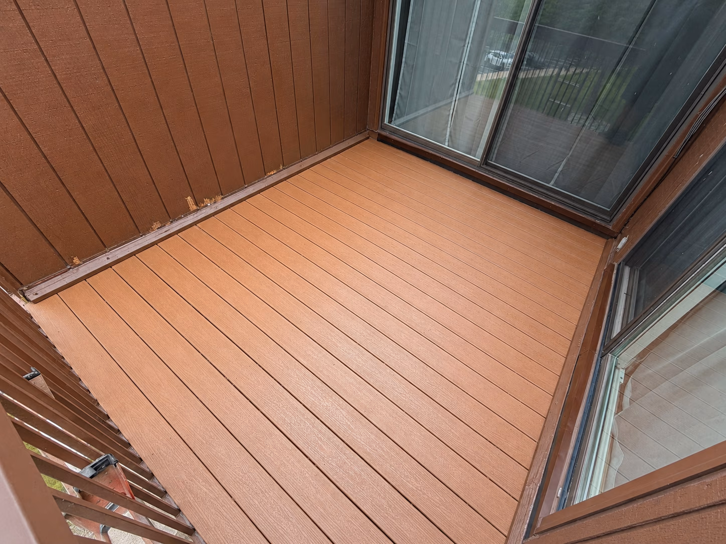 Empty small balcony with brown composite wood flooring and brown wooden walls with sliding glass doors.