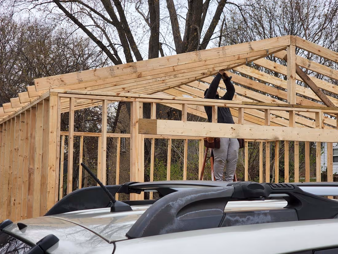 Person on ladder constructing the wooden frame of a building roof on a cloudy day.