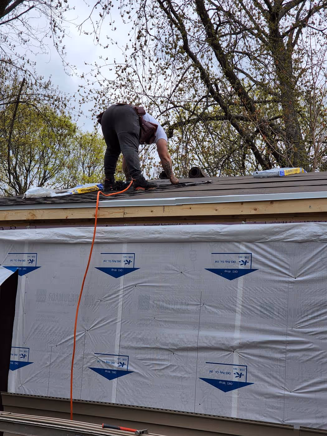 Worker installing roofing materials on the roof of a house under construction with trees in the background.