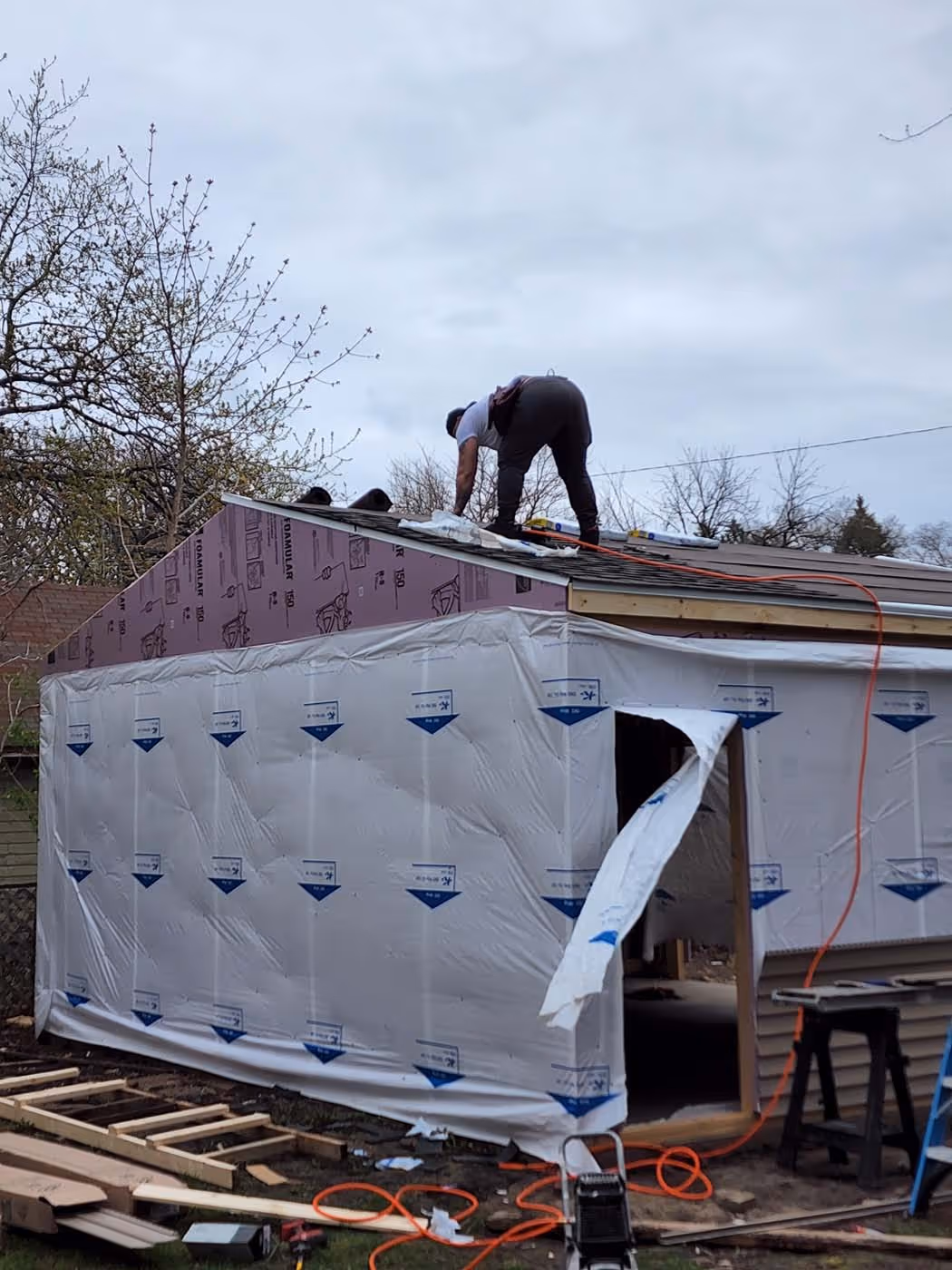 Construction worker installing roofing shingles on a partially built small building covered with white plastic sheeting on the walls.