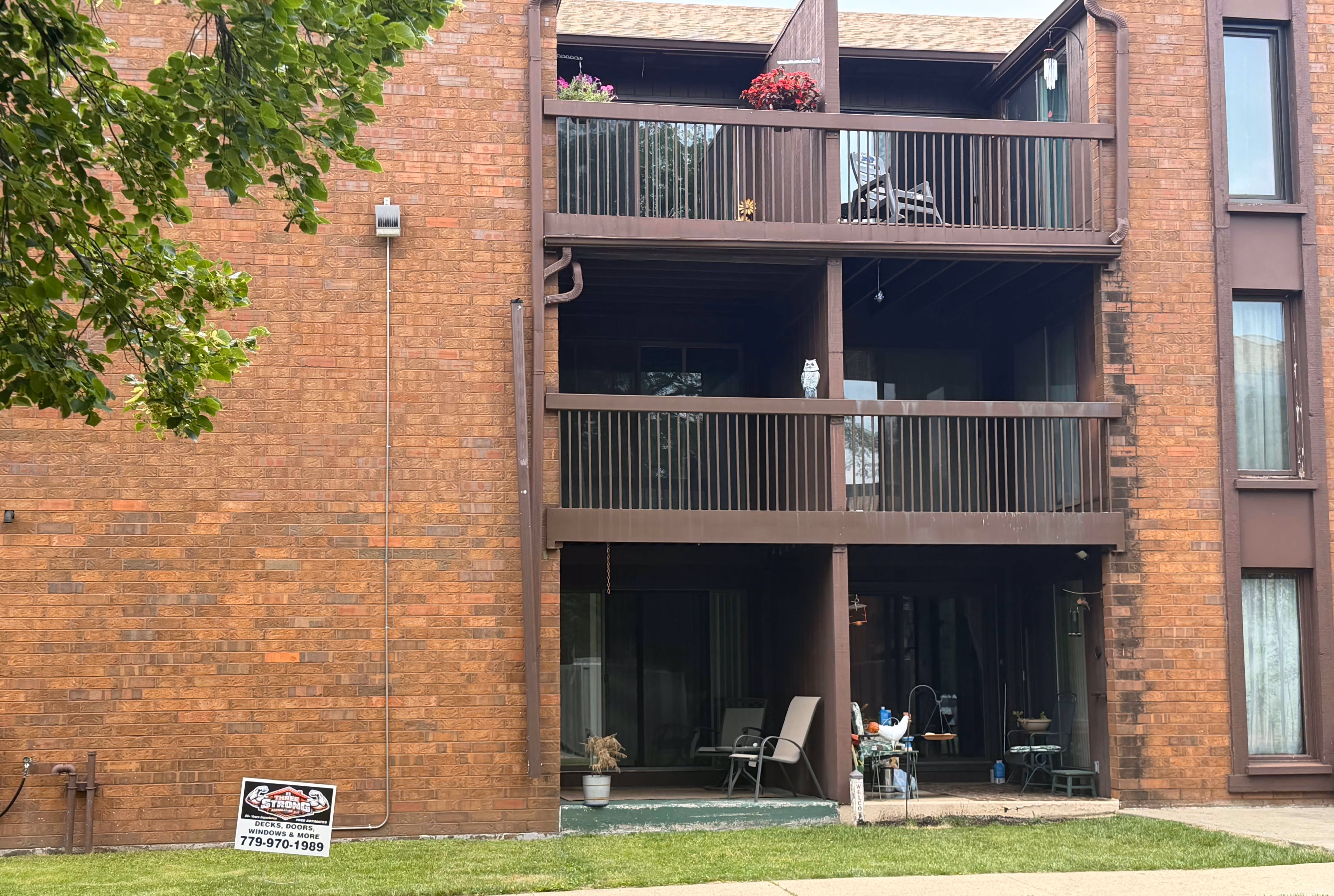 Brick apartment building with three balconies, outdoor chairs, plants, and a lawn sign in front.