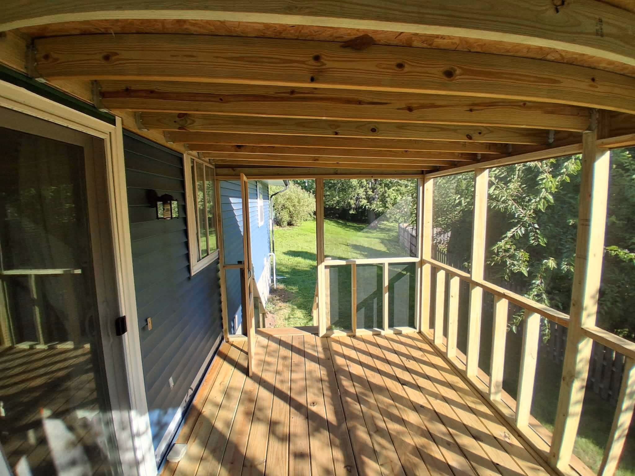 Sunlit screened porch with wooden floor and ceiling attached to blue house, overlooking green yard.