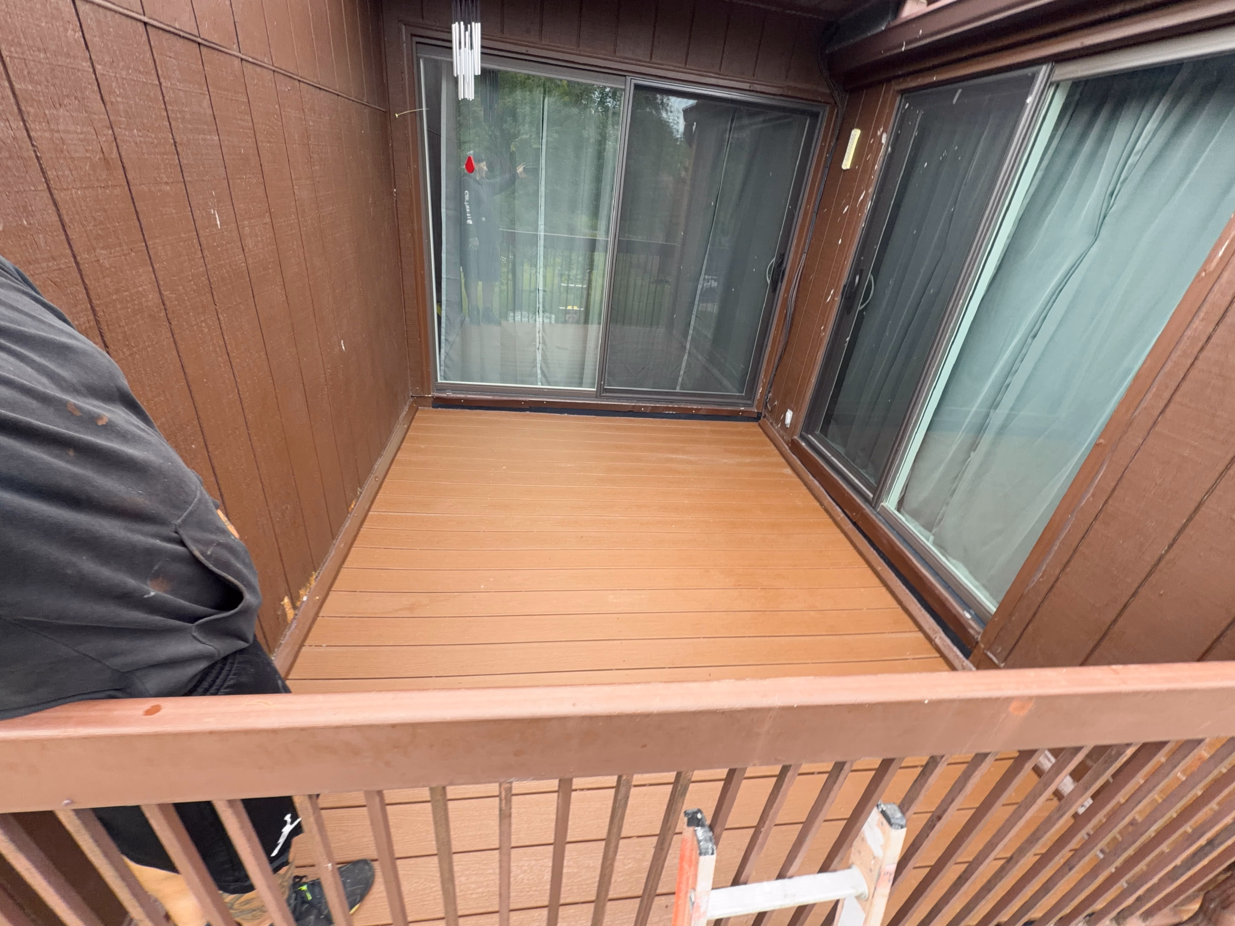 Small brown wooden balcony with sliding glass doors and a ladder leaning on railing.