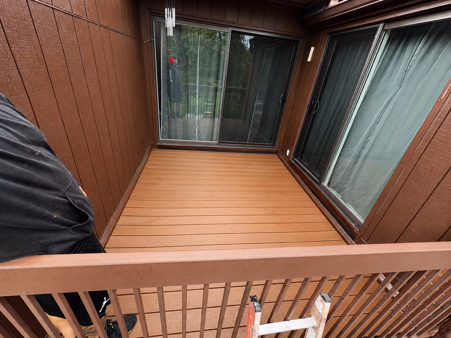 Empty wooden balcony with brown railing and sliding glass doors, reflecting a person and trees.
