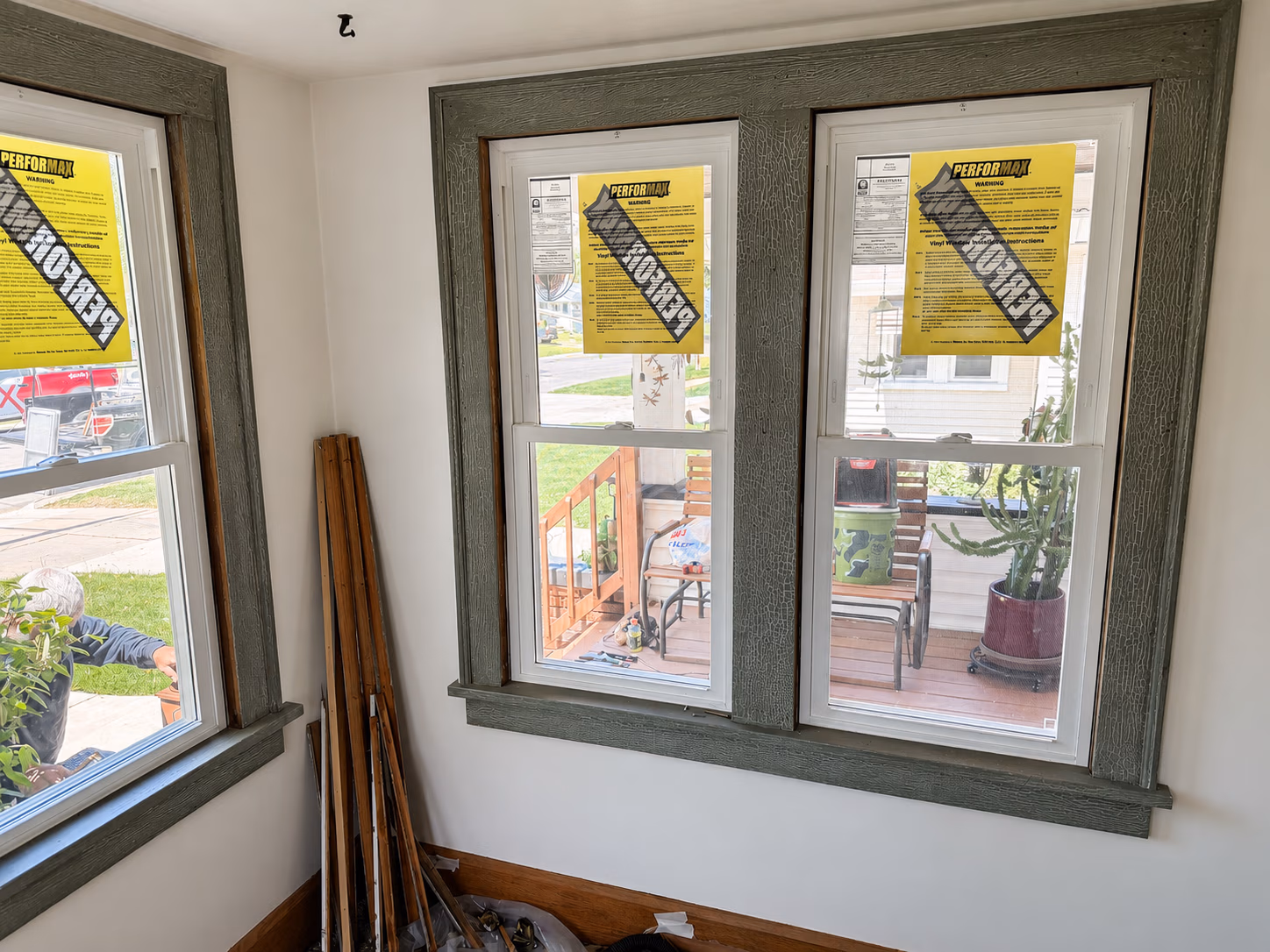 Three windows with green frames and yellow warning stickers overlooking a porch with plants and chairs.
