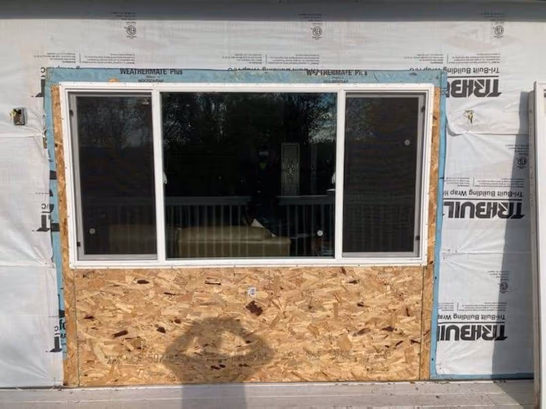Newly installed three-panel sliding window in construction wall covered with house wrap and plywood on lower section.