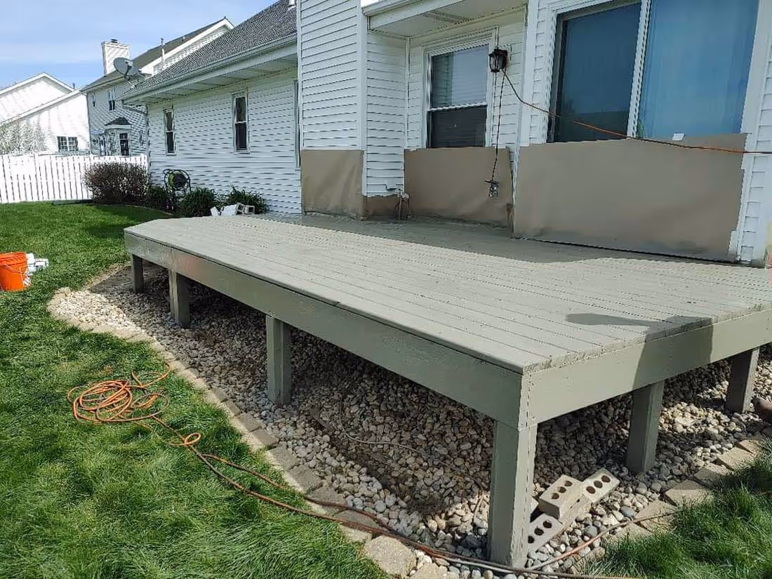 Wooden outdoor deck painted beige with gravel underneath, attached to a white house with a green lawn and orange extension cords on the grass.