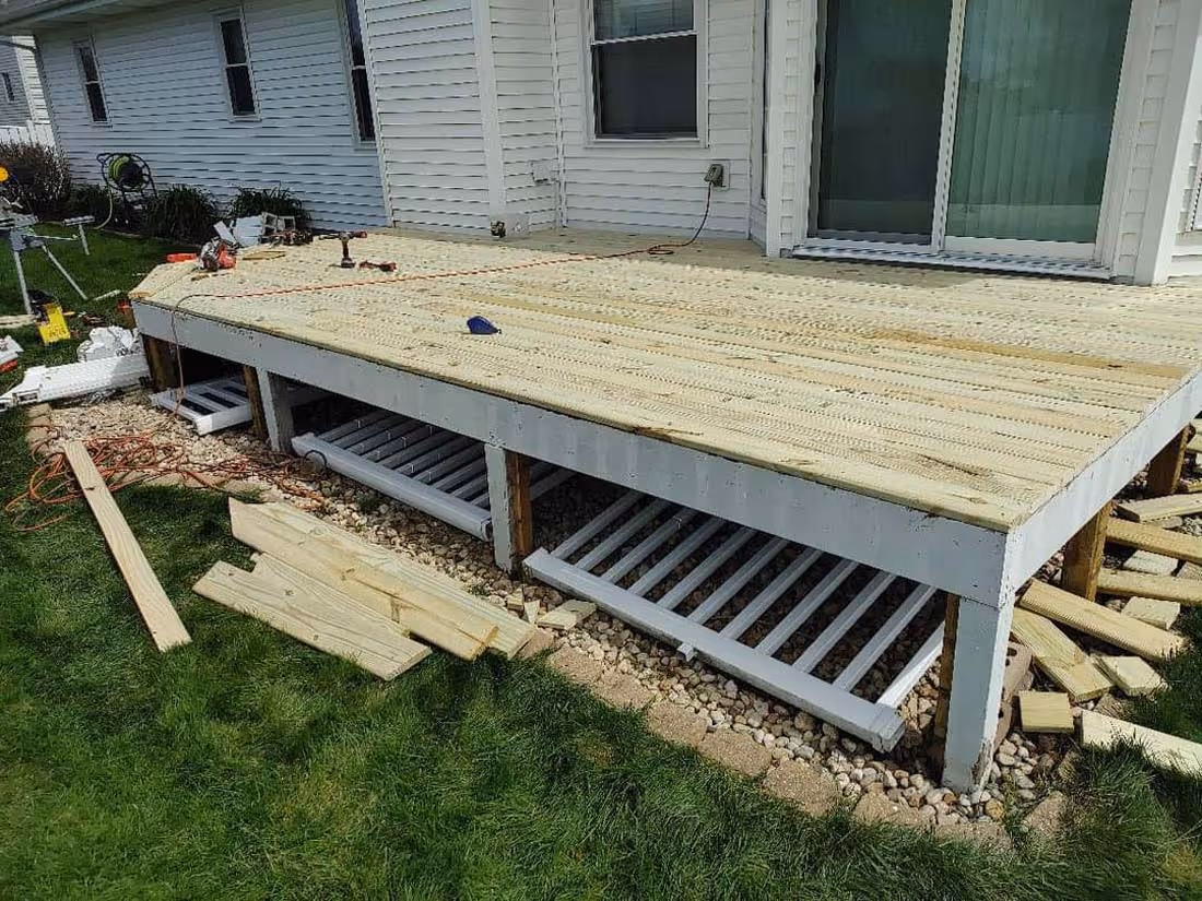 Wooden deck under construction attached to a white house with building materials and tools nearby on the grass.