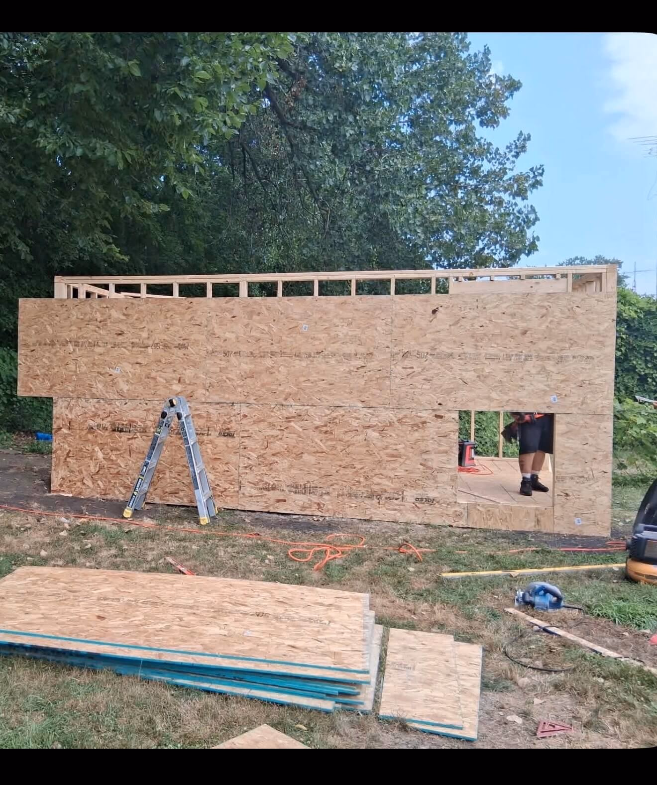 Half-built wooden structure with plywood walls, a ladder, and construction materials on grass.