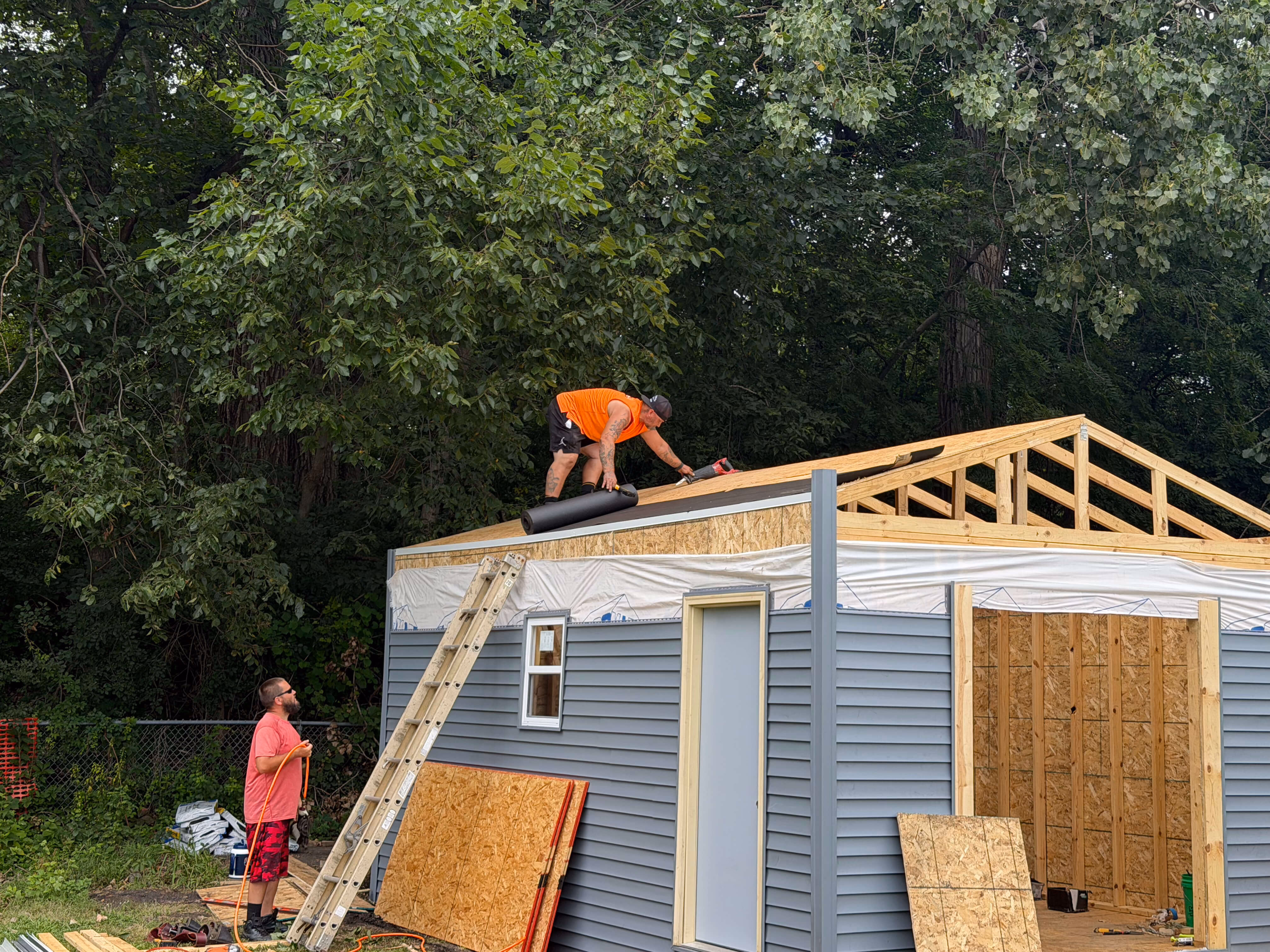 Two men building a small house; one on ladder holding cord, one on roof rolling roofing material.