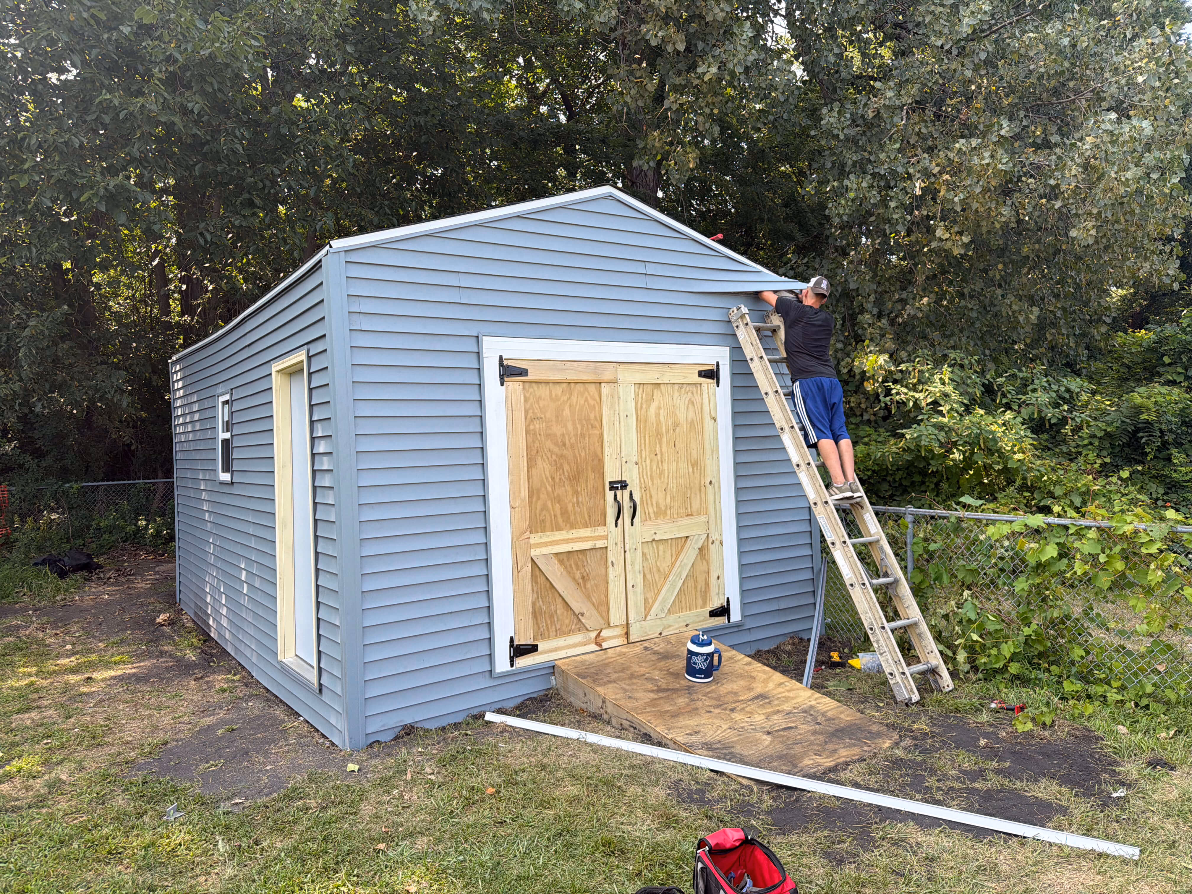 Man on ladder working on gray shed with wooden double doors and ramp in a grassy yard.