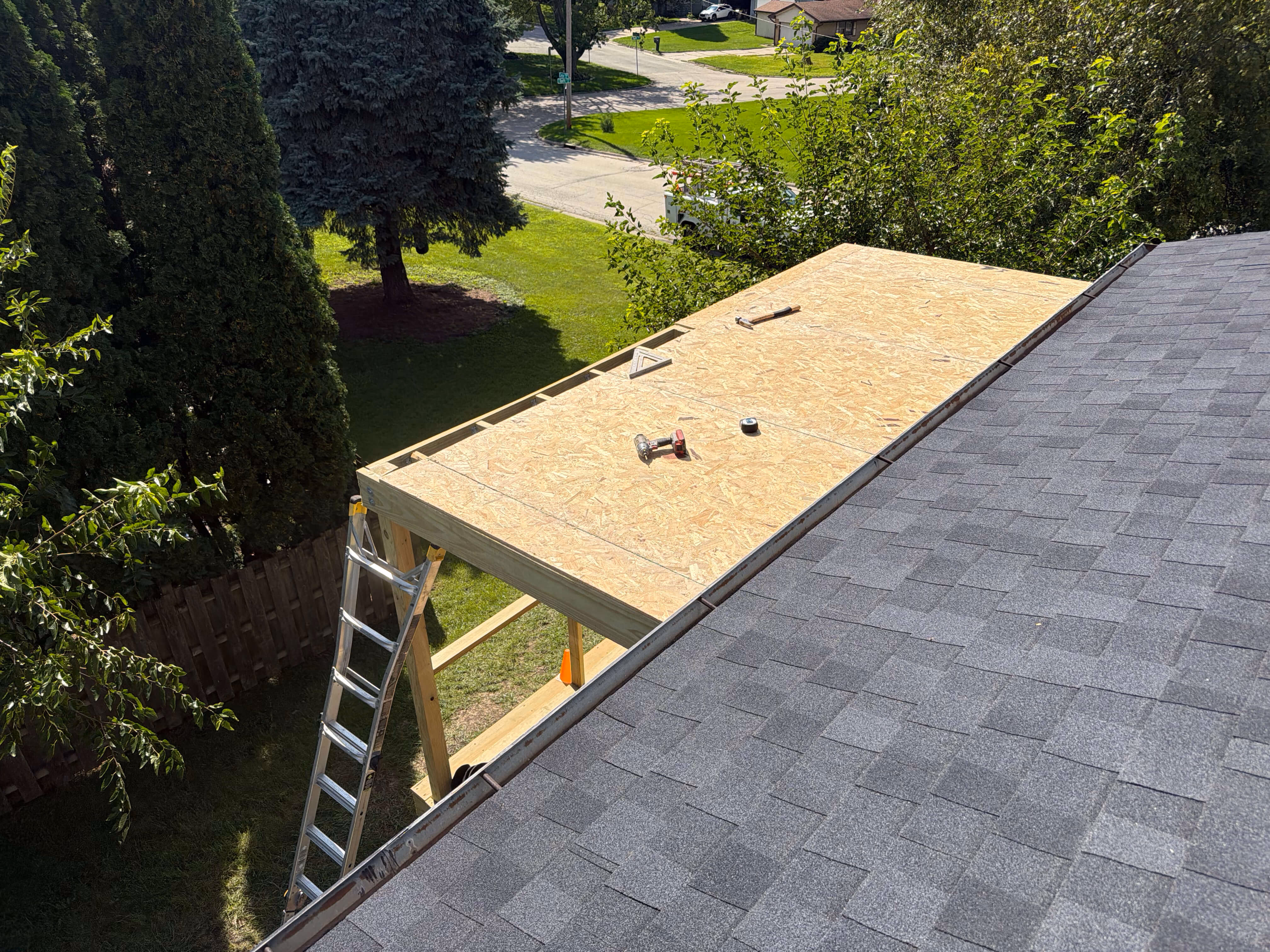 Partially constructed wooden roof extension with tools on it, next to an existing shingled roof.