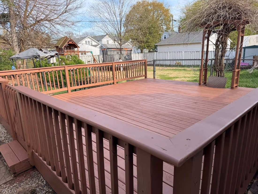 Spacious wooden deck with railing overlooking a backyard with trees, a swing set, and a gazebo.