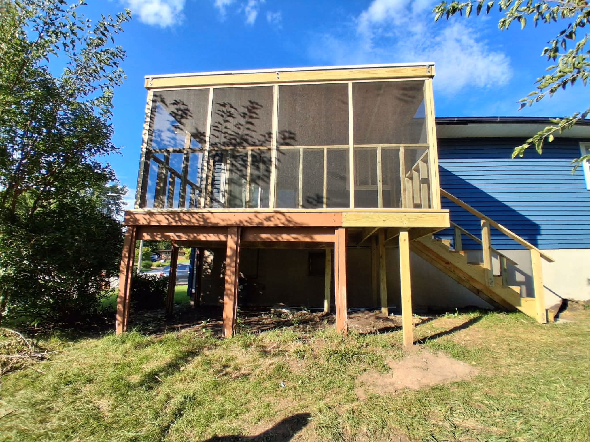 Wooden screened porch with stairs attached to a blue house under a clear blue sky.