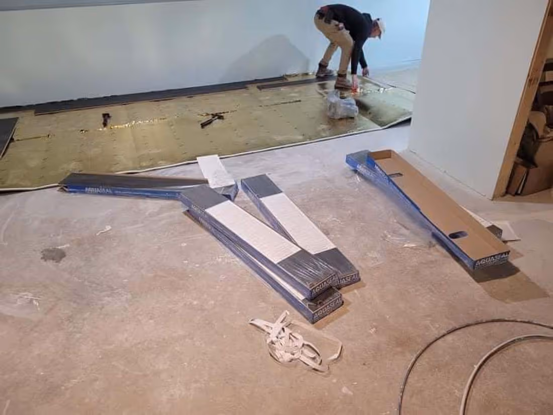 Person installing flooring over golden underlayment with packaged floorboards on bare concrete floor.