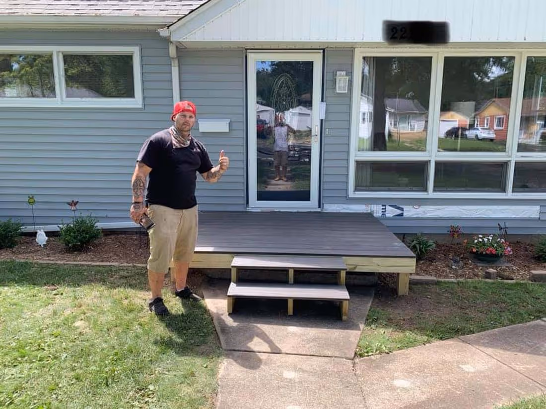 Man with tattoos wearing a red cap and black shirt giving a thumbs up while standing next to a newly built small wooden front porch with two steps in front of a gray house.