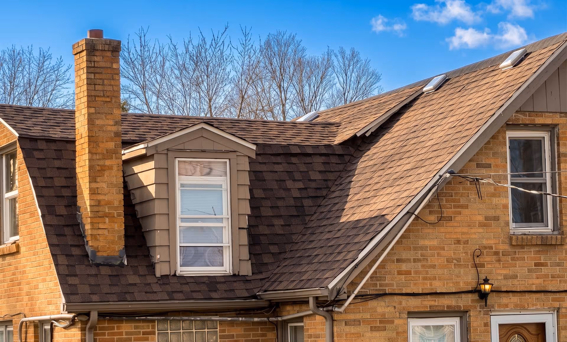 House roof with brown shingles, a brick chimney, dormer window, skylights, and bare trees in the background under a blue sky.