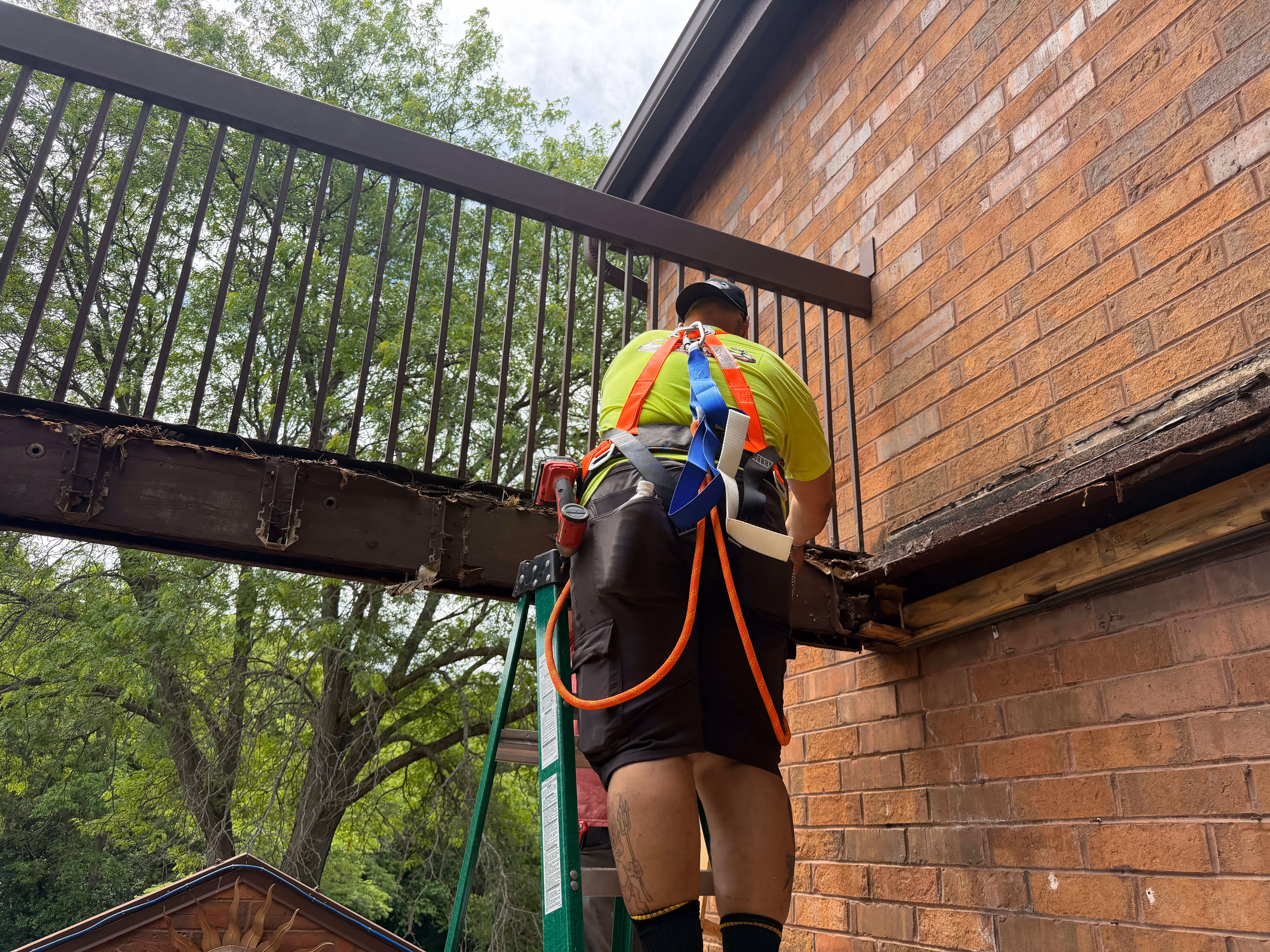 Worker wearing a safety harness standing on a ladder repairing a damaged wood balcony railing.