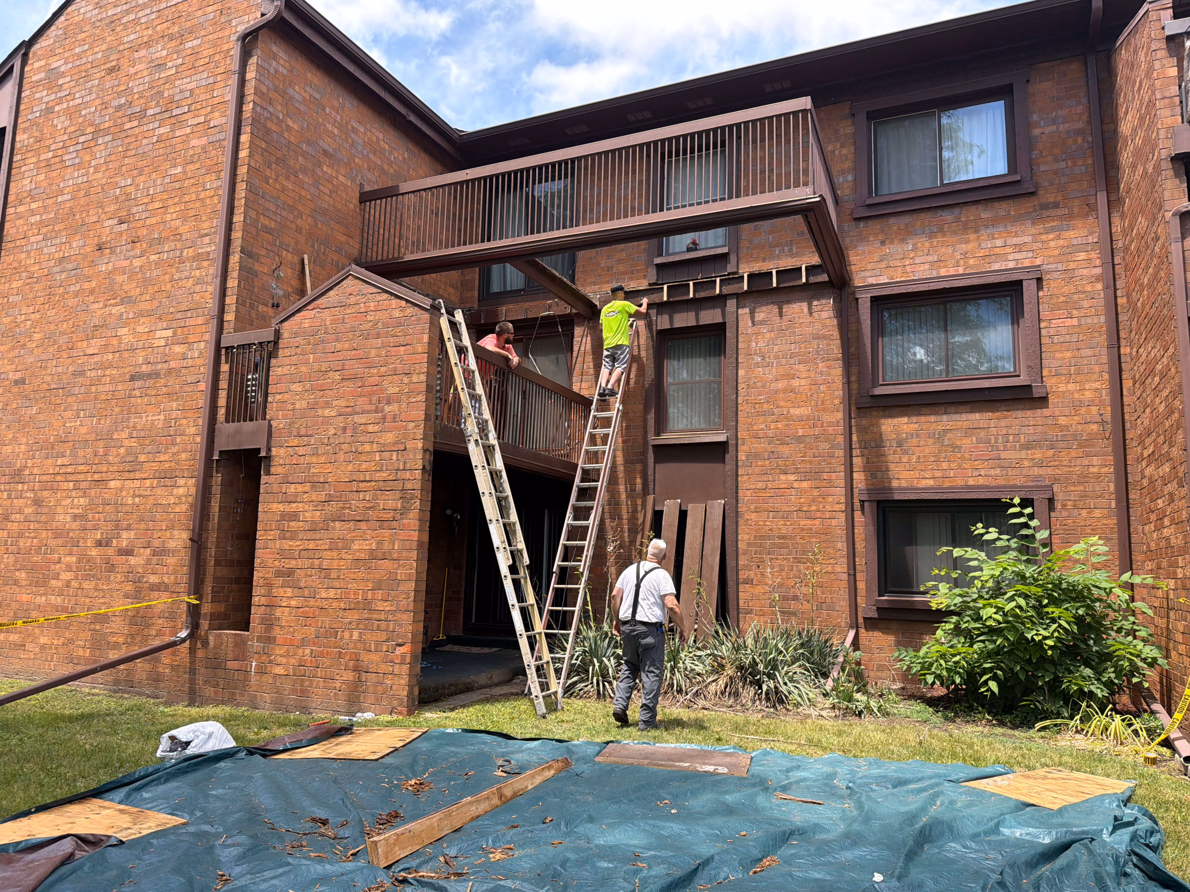 Three workers using ladders to repair or build a balcony on a brick apartment building exterior.