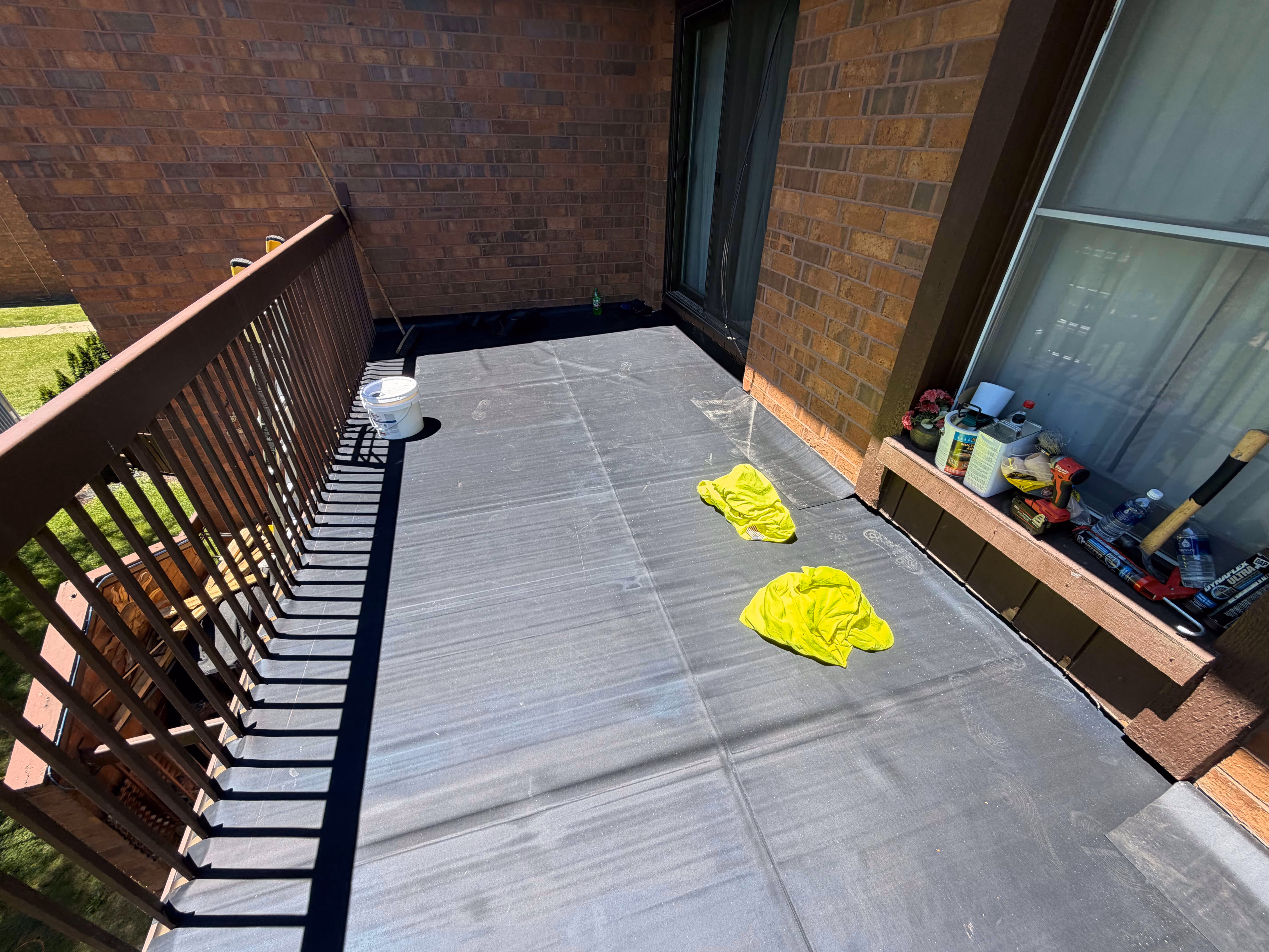 Balcony with brown railing, black floor covering, two yellow cloths, white bucket, and tools on a ledge.
