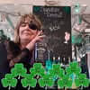 Person with glasses smiling behind a table decorated with several green shamrocks and a chalkboard with writing in the background.