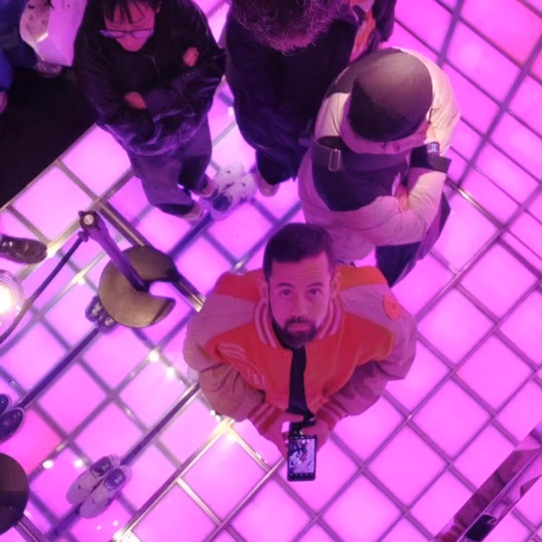 Overhead view of a group of people standing on a glowing purple grid floor, one man in a red and beige jacket looking up holding a smartphone.