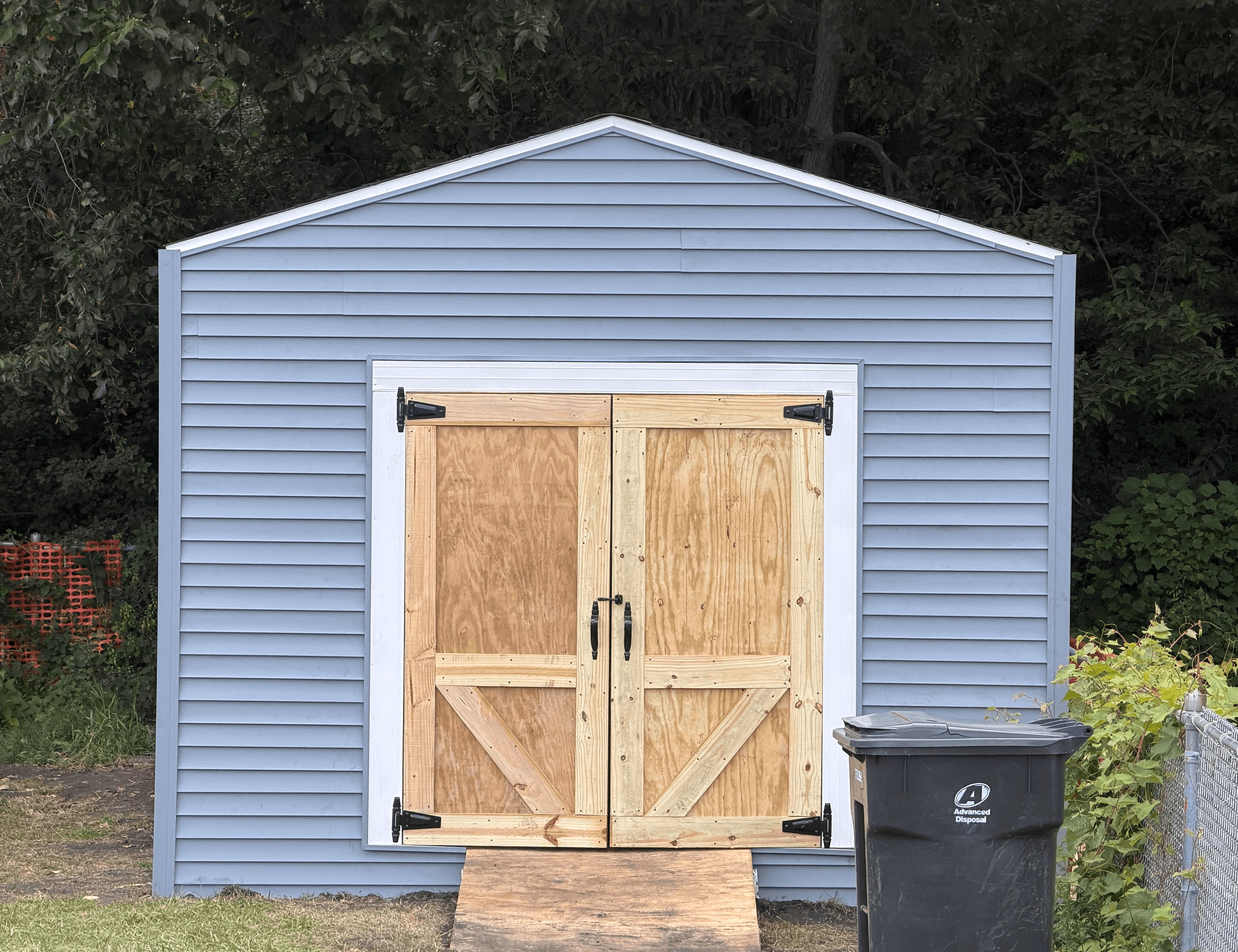 Blue shed with double wooden doors and a ramp, next to a black Advanced Disposal trash bin.