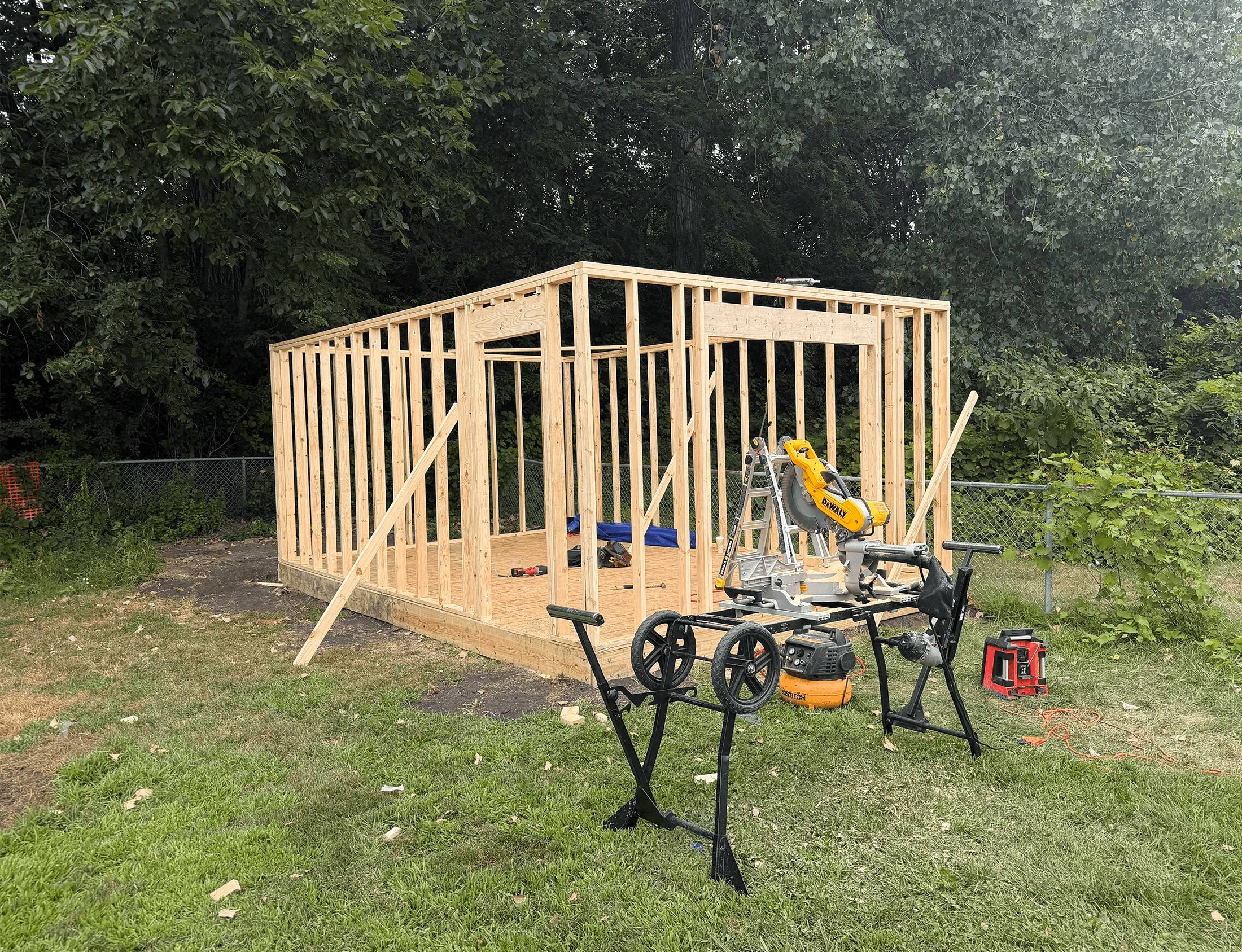 Wooden frame structure under construction with a yellow DeWalt miter saw on a stand in a grassy yard.