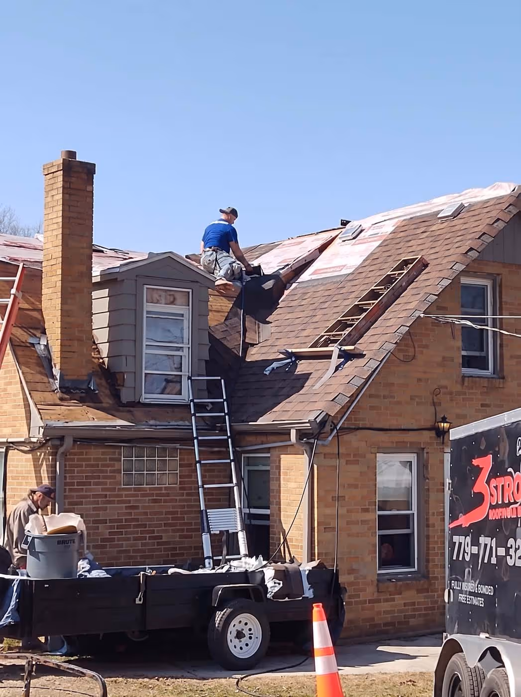 Construction workers repairing the roof of a brick house with ladders and roofing materials on a clear day.