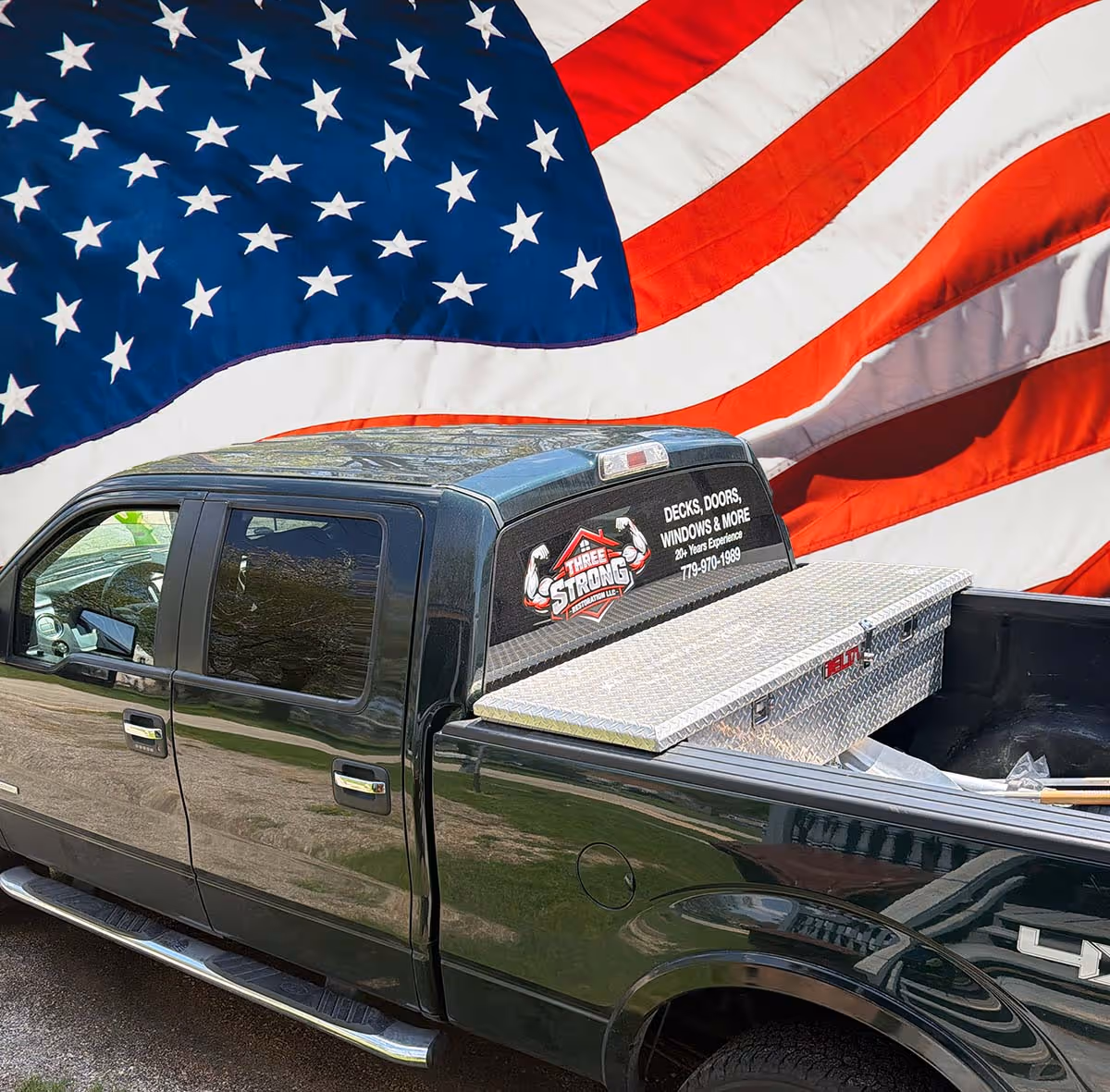 Black pickup truck with a silver toolbox in the bed and an American flag backdrop, featuring a decal advertising Three Strong Restoration LLC specializing in decks, doors, and windows.