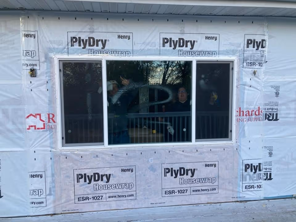 A white-framed sliding window installed in a house under construction with PlyDry Housewrap visible around it and two people working inside.
